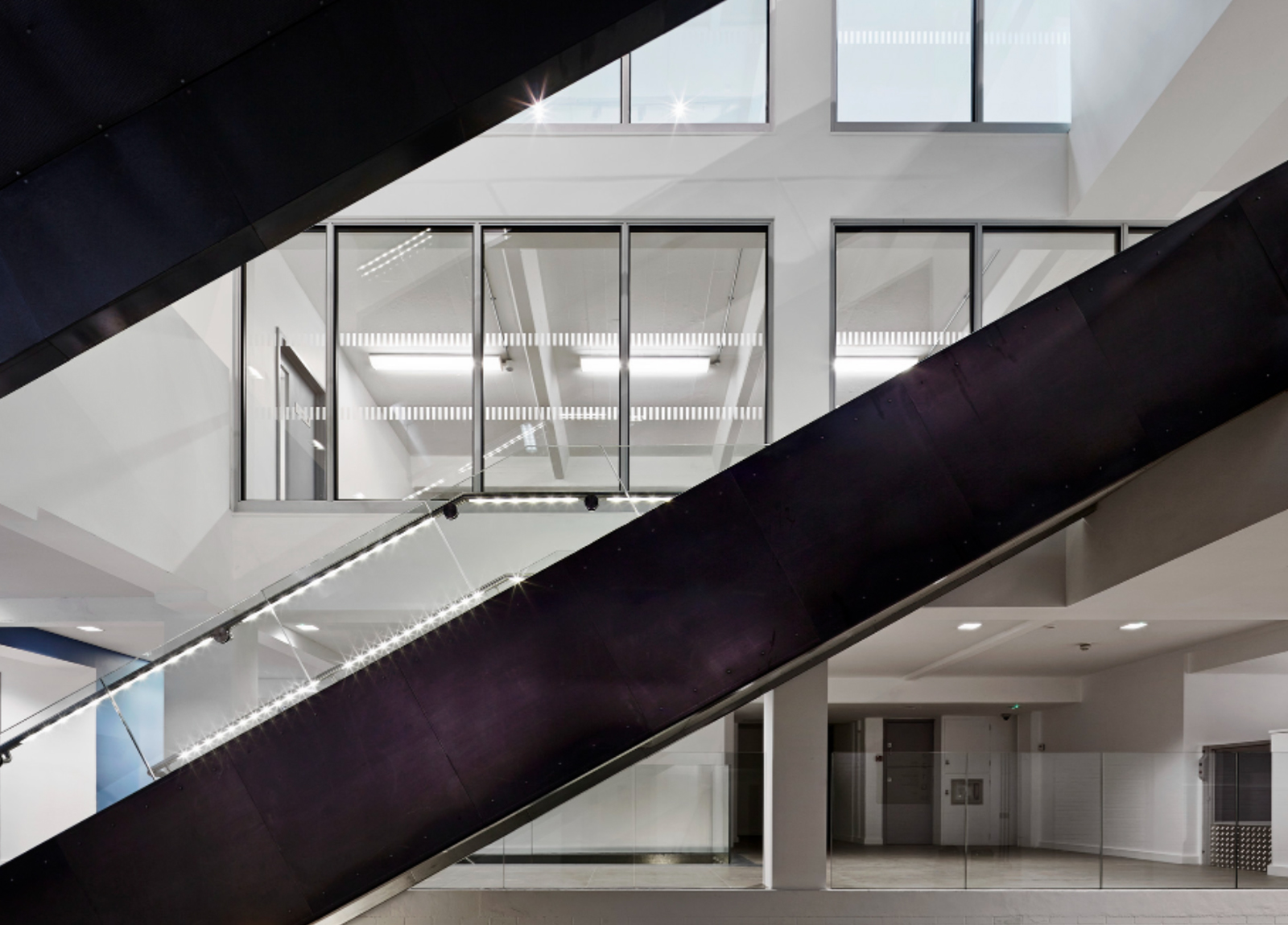 Modern building interior with large glass windows and two dark escalators crossing diagonally in front; bright, minimalistic design with white walls and fluorescent lighting.