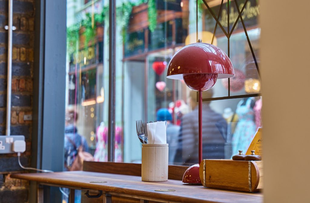 Photo of a window seat in a restaurant. A small, domed red lamp sits on a wooden table next to a beige pot holding cutlery and napkins.