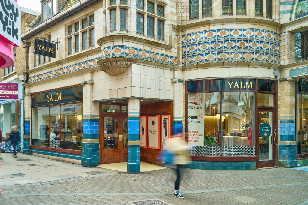 Photo of the outside of Yalm. An old, white-brick shop, with a sign of gold lettering on a black background. People are pictured blurred as they walk past.