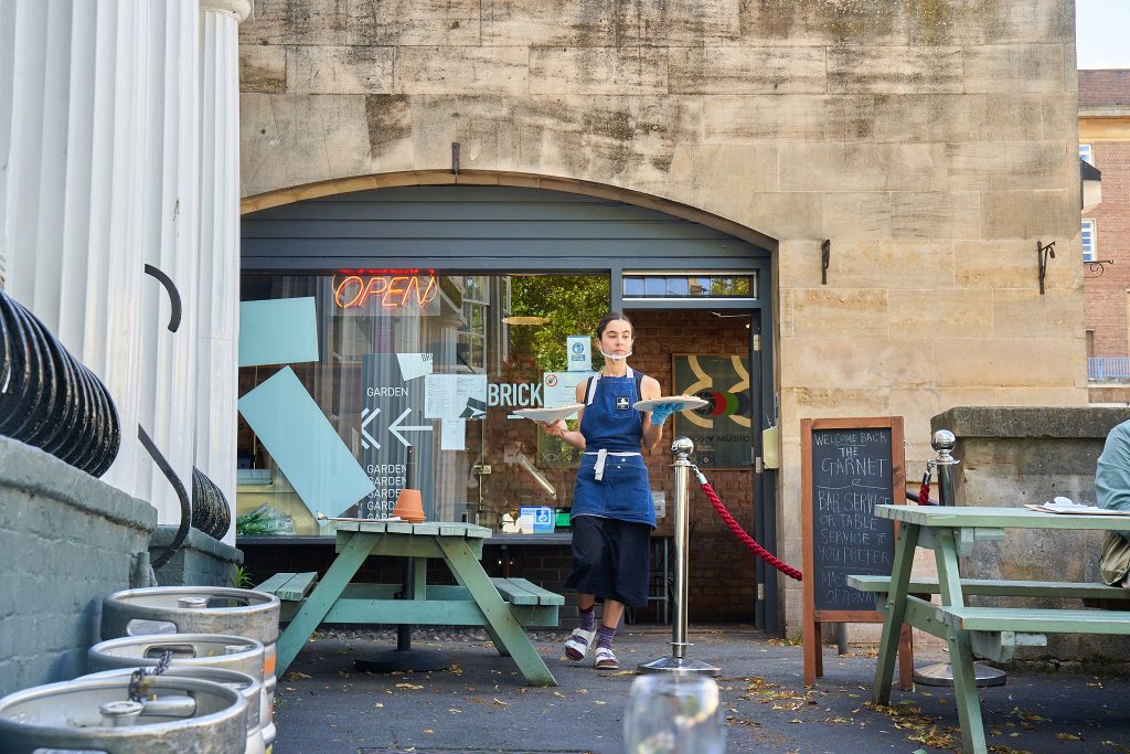 Exterior shot of Brick Pizza - a person in a blue apron is walking out of the shop holding a pizza in each hand.