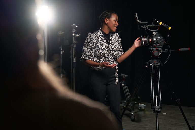 Alumni Tanicha wears a black and white blouse, standing next to a film camera. A light is set up on a tripod behind them.