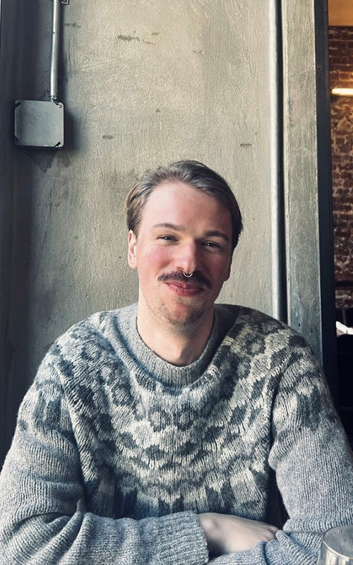 Photo of Animation alumni August Abrahamsson. August is smiling at the camera, wearing a grey jumper and sitting with his arms folded. There is a grey wall behind him and a brick wall can be seen through a door to his left.