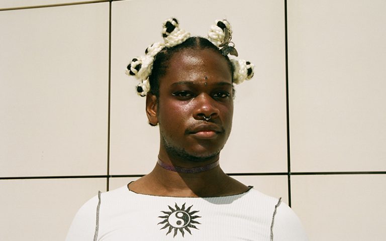 Portrait photo of musician Shamir. Shamir is dressed in white, they have several piercings and a metal butterfly in their hair.