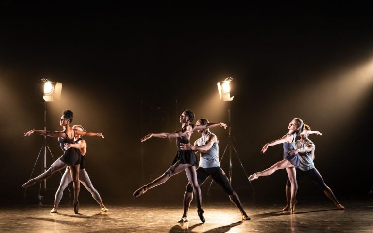 Photo of six ballet dancers in pairs, caught mid-pose. The stage is dark apart from two lights.