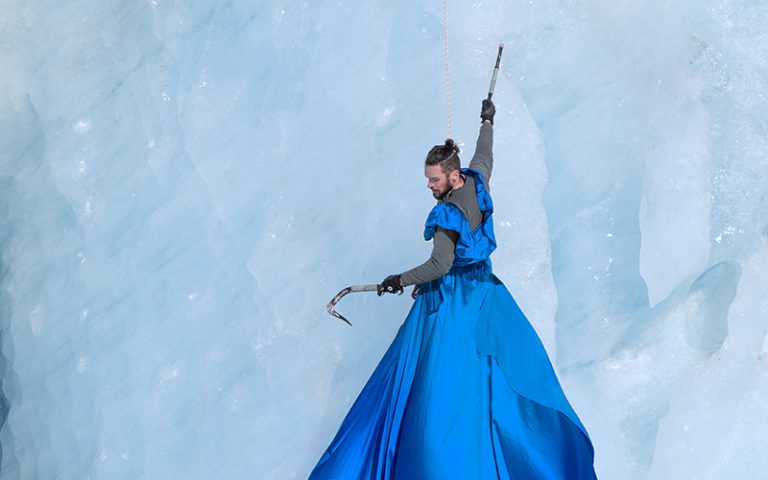 'The Ice Climber' Photograph by Cat Gundry-Beck. A climber hangs from a wall of ice from one arm. He wears a blue dress which trails down far below him.