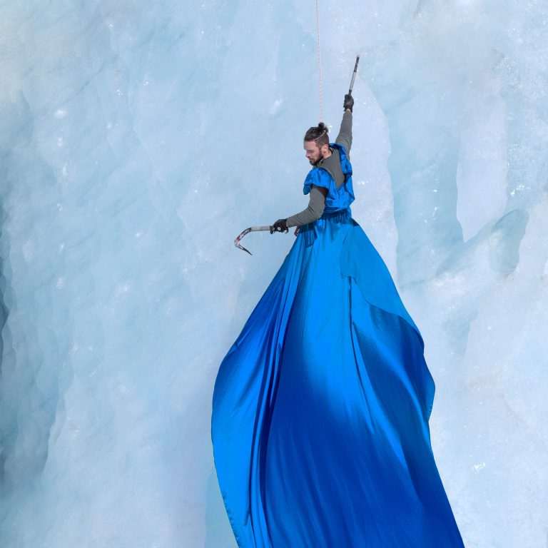 'The Ice Climber' Photograph by Cat Gundry-Beck. A climber hangs from a wall of ice from one arm. He wears a blue dress which trails down far below him.