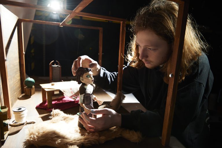 A person with long, ginger hair and dark green jumper is adjusting a stop motion maquette. The maquette is illuminated by a spotlight.