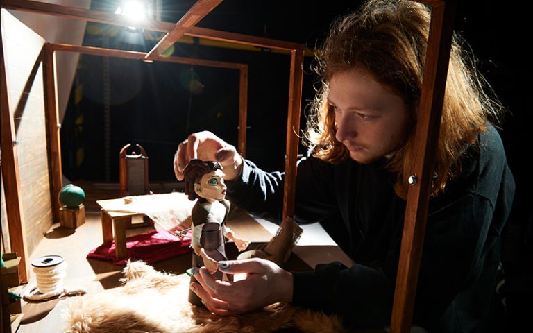 A person with long, ginger hair and dark green jumper is adjusting a stop motion maquette. The maquette is illuminated by a spotlight.