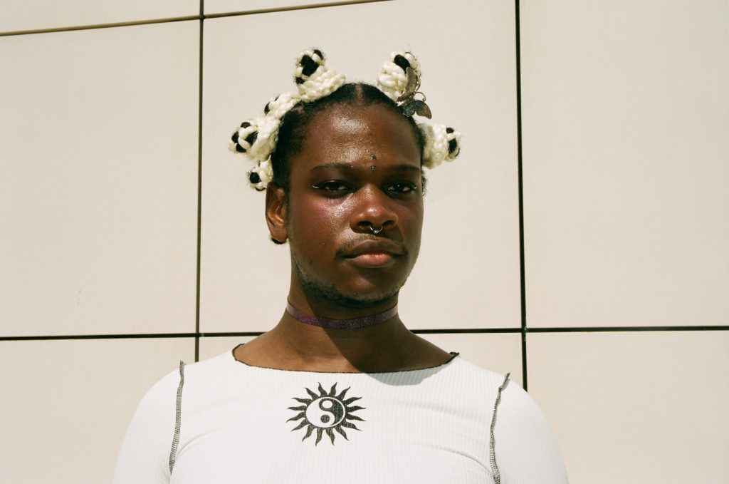 Portrait photo of musician Shamir. Shamir is dressed in white, they have several piercings and a metal butterfly in their hair.