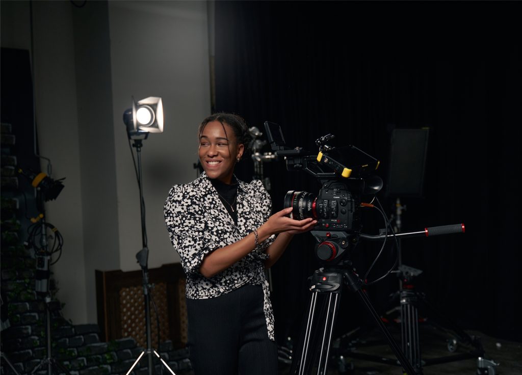 Alumni Tanicha wears a black and white blouse, standing next to a film camera. A light is set up on a tripod behind them.