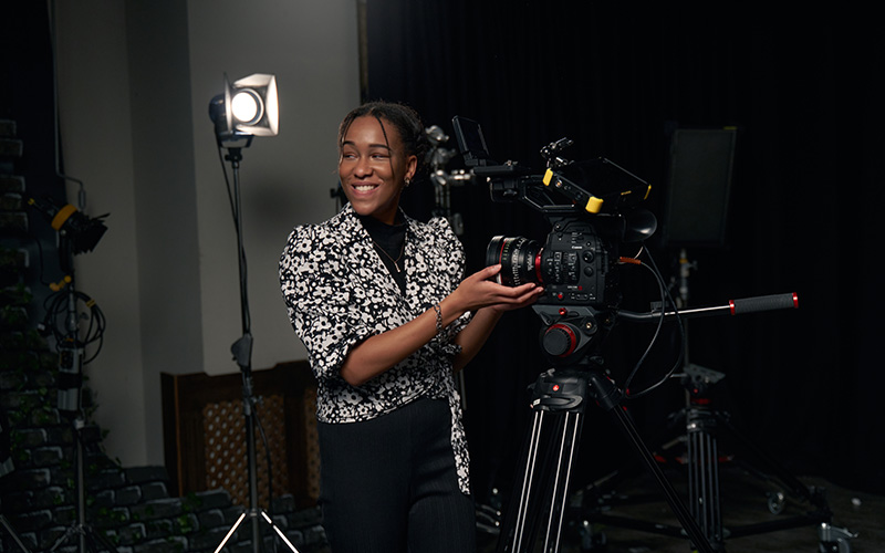 Alumni Tanicha wears a black and white blouse, standing next to a film camera. A light is set up on a tripod behind them.