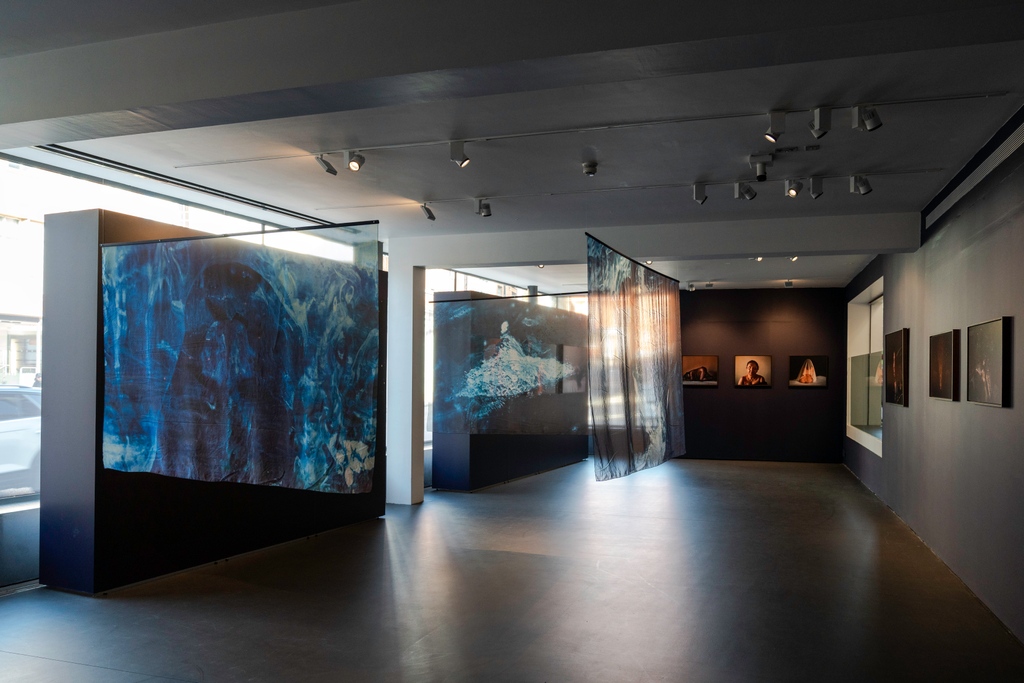 Photograph of East Gallery exhibition 'Fireflies' by Poulomi Basu. The room is dark with, several large screens and drapes hanging from the ceiling.