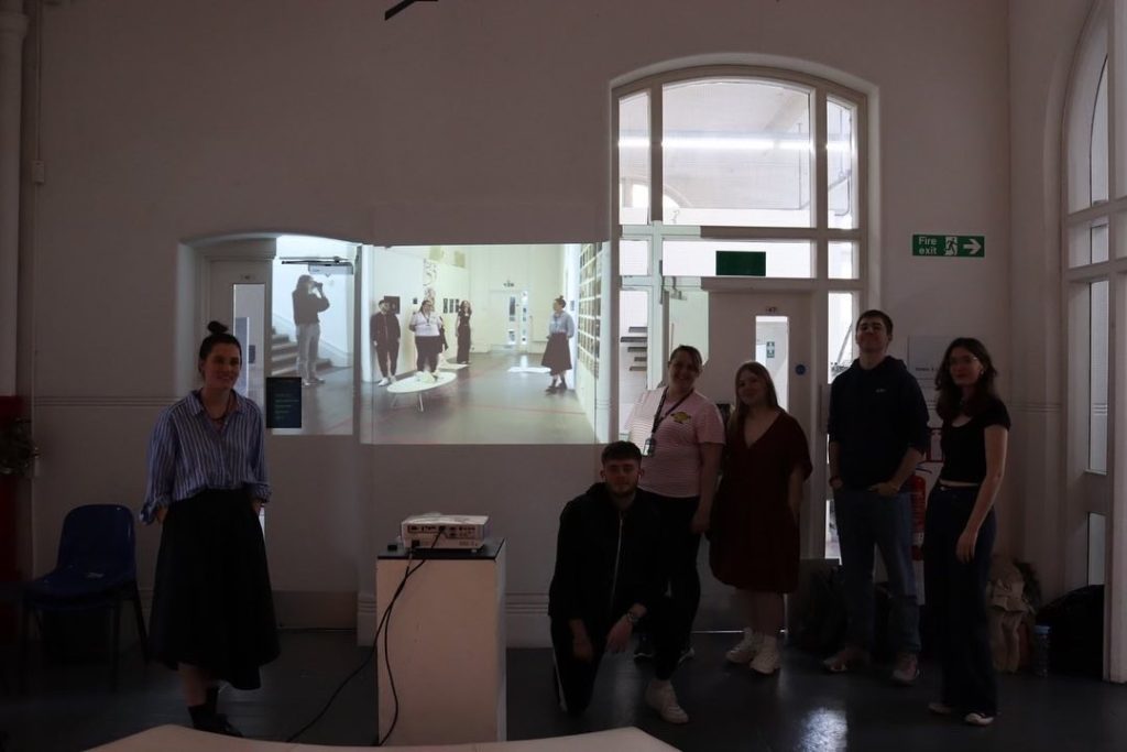 People have posed for a photo around a projector screen. On the screen can be seen an image of the photographed group taken from a different angle.