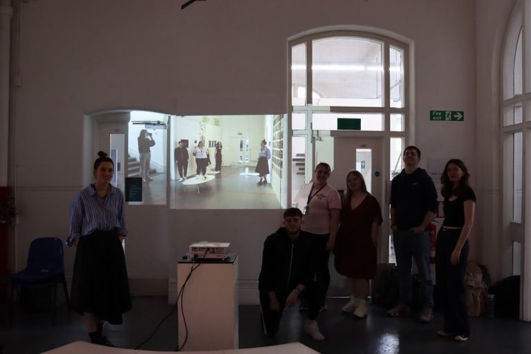 People have posed for a photo around a projector screen. On the screen can be seen an image of the photographed group taken from a different angle.