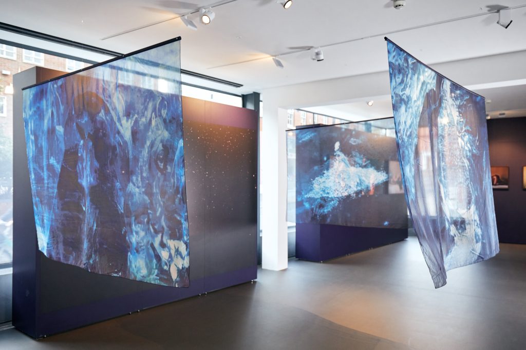 Three translucent, landscape drapes are hung in a rectangle formation. They have blue patterns printed on them, suggestive of crashing waves.