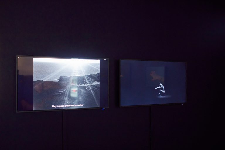 Two screens are mounted next to each other in a dark room. The left screen shows a black and white cliff looking out to sea, the other shows a dancer in a black room.