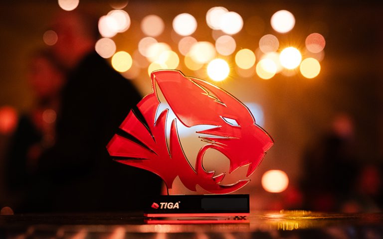 A red and white trophy shaped like a stylised tiger head is displayed prominently against a background of blurred, warm lights. The trophy has the word "TIGA" on its base.