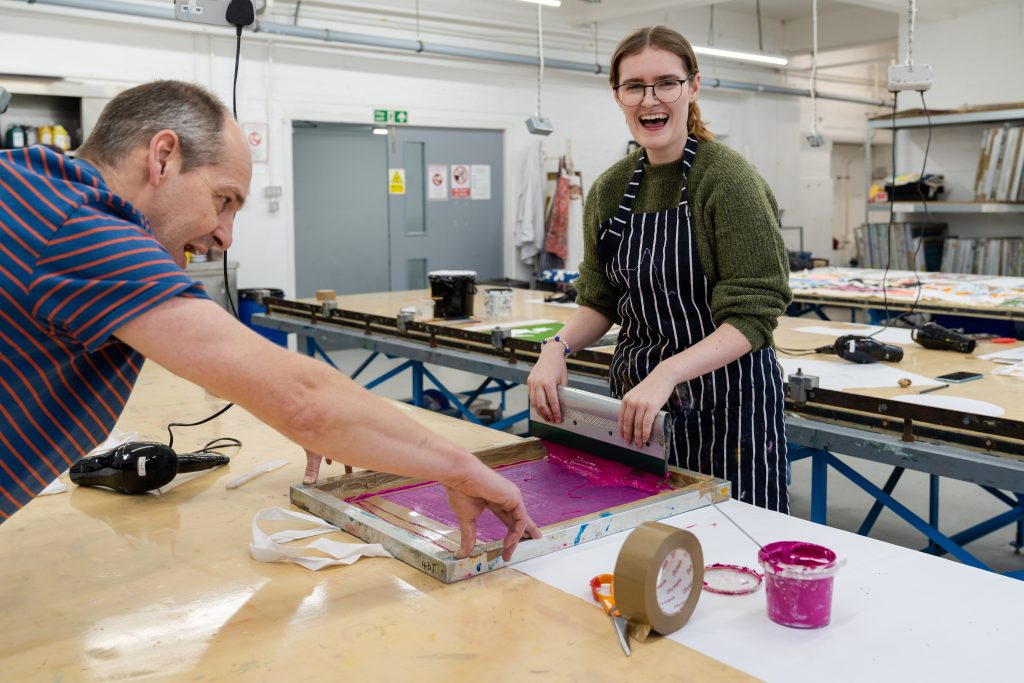 A student screen printing onto a tote bag in the Textile Design department. The student is smiling and looking at the camera.