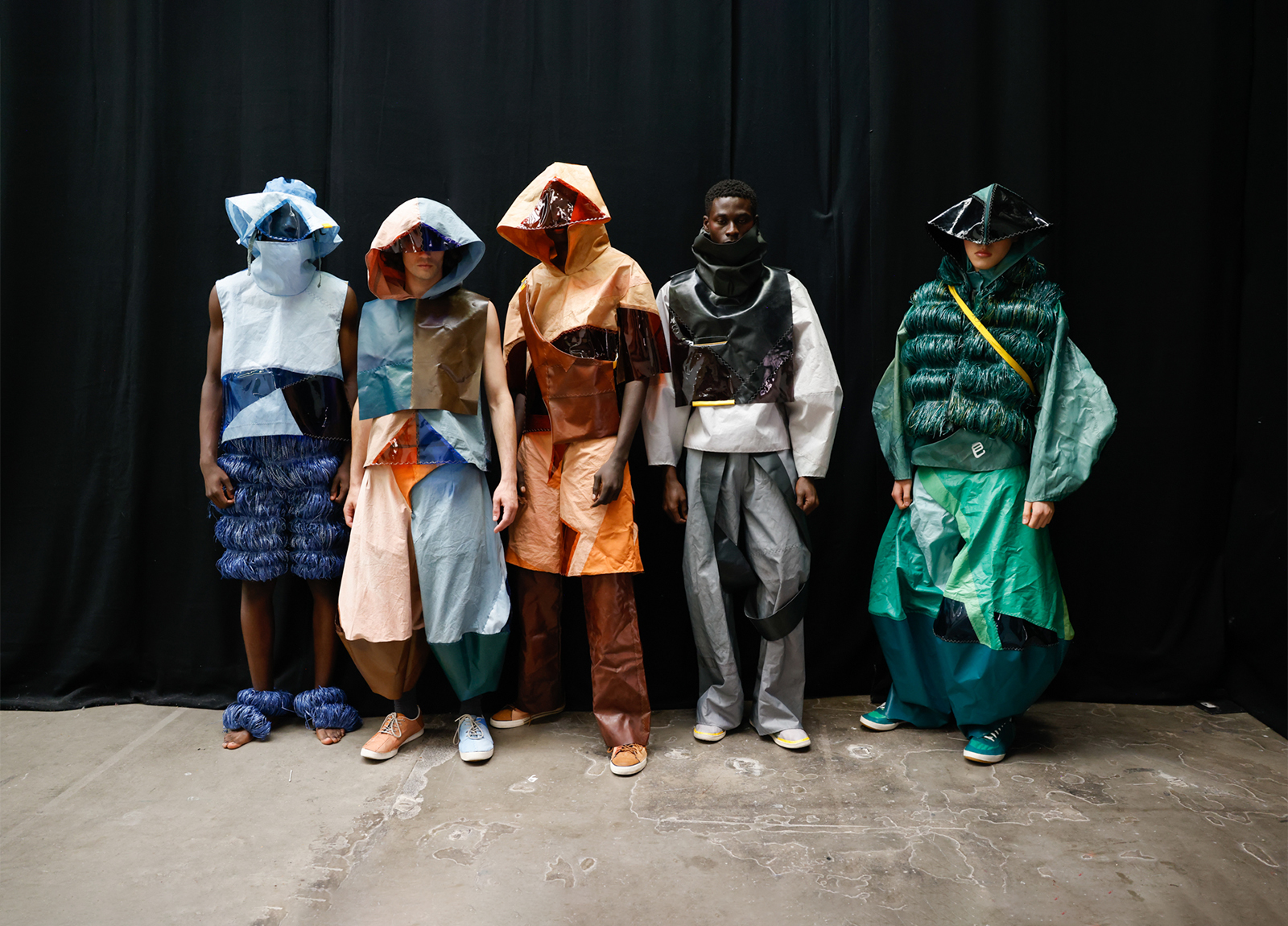 Student work by Ella Taylor for LaceyAyles. 5 models stand in a row in front of a black curtain, wearing garments with exaggerated shapes