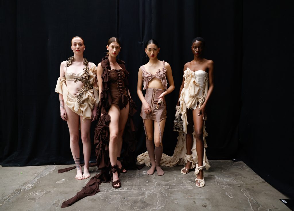 Student work by Ciara Spencer for LaceyAyles. 4 models wearing garments in shades of khaki and brown in front of a black curtain