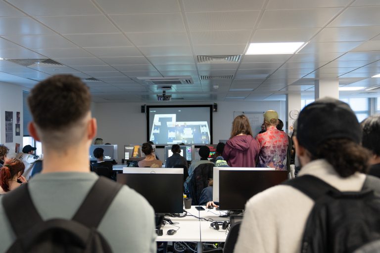 Two people are standing with their back to the camera. Over their shoulders a group of people can be seen watching a screen on the wall. A game is being projected onto the screen.