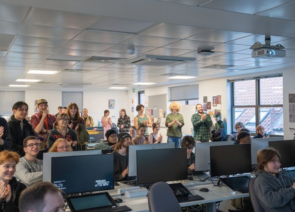 A large group of people are in a computer room looking at a screen. Many are applauding and smiling.