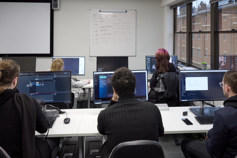 A group of people working at computers, taken from behind so the screens can be seen. On the screen people are working on coding a game.