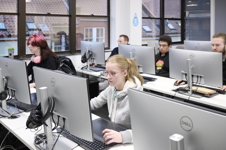 A group of people are working at computers; we can see their faces concentrating on the monitors and the back of the screens.