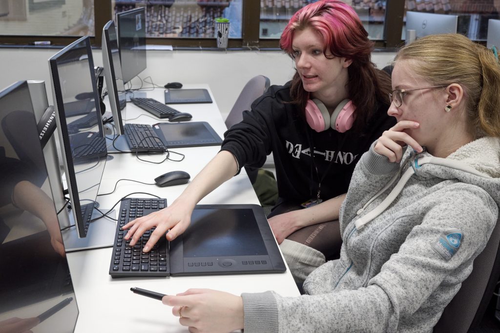 Two people are working together at a computer. One person looks puzzled holding their chin, whilst the other explains something on the screen.