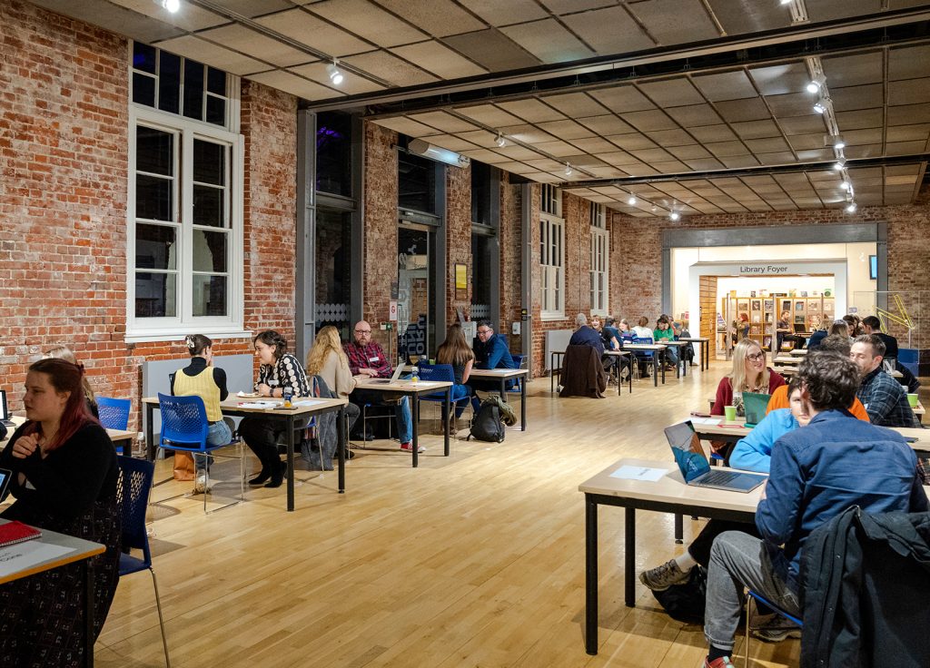 A photo from The Big Book Crit 2024 in Duke Street foyer. The image is of the room filled with students and industry guests sat down at tables looking through the students work and or portfolios.