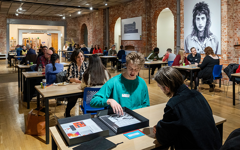 A photo from The Big Book Crit 2024 in Duke Street foyer. The image is of the room filled with students and industry guests sat down at tables looking through the students work and or portfolios.