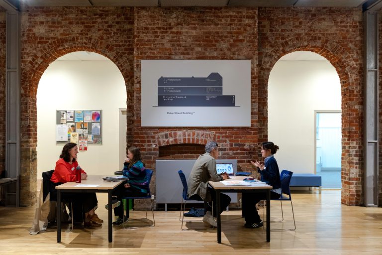 A photo from The Big Book Crit 2024 in Duke Street foyer. The image is of students and industry guests sat at tables reviewing and discussing the students work and or portfolio.