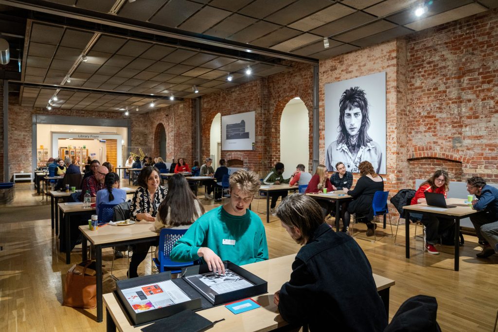 A photo from The Big Book Crit 2024 in Duke Street foyer. The image is of the room filled with students and industry guests sat down at tables looking through the students work and or portfolios.