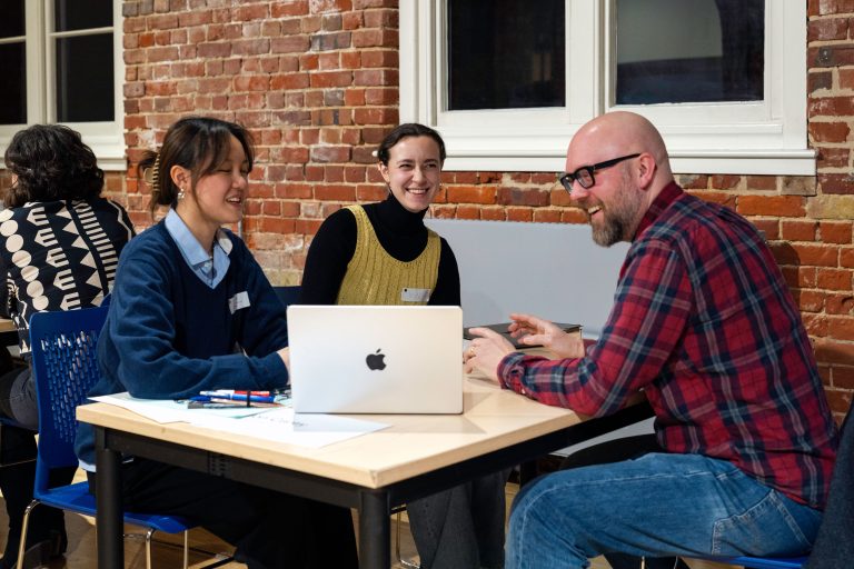 A photo from The Big Book Crit 2024 in Duke Street foyer. The image is of two students and an industry guest sat down at a table reviewing the students work and or portfolios on a laptop.