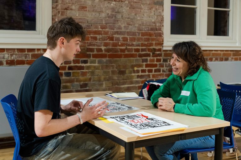 A photo from The Big Book Crit 2024 in Duke Street foyer. The image is of a student and an industry guest sat down at a table reviewing the students work and or portfolio. The work is spread out across the table where it is physically being interacted with by the student.