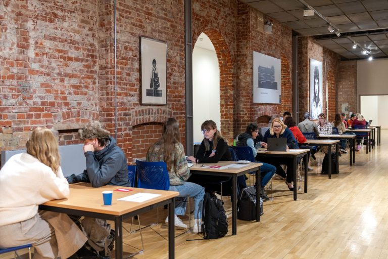 A photo from The Big Book Crit 2024 in Duke Street foyer. The image is of the room filled with students and industry guests sat down at tables looking through the students work and or portfolios.