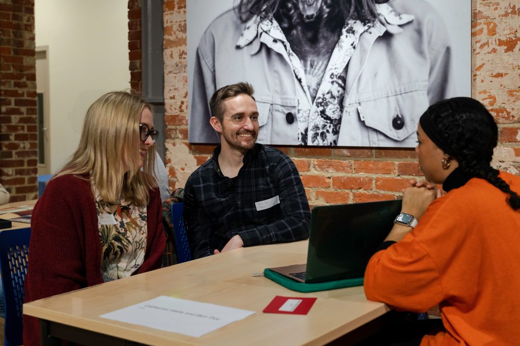 A photo from The Big Book Crit 2024 in Duke Street foyer. The image is of a student and two industry guests sat down at a table reviewing the students work and or portfolio on a laptop.