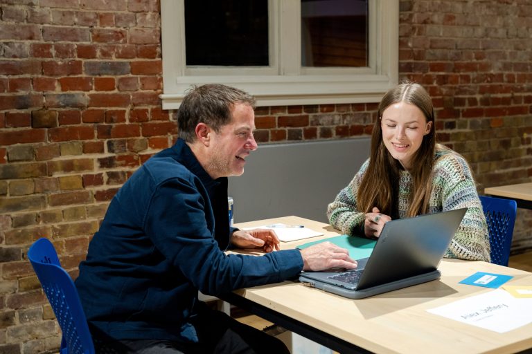 A photo from The Big Book Crit 2024 in Duke Street foyer. The image is of a student and an industry guest sat down at a table reviewing the students work and or portfolio on a laptop.