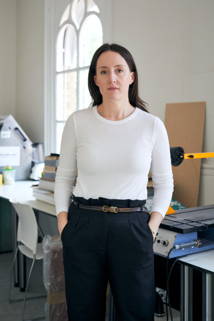 A woman with long dark hair wearing a white long-sleeve top and black pants stands with her hands in her pockets. She is in an office or studio with tables, stacked papers, books, and equipment in the background. The room has a large window letting in natural light.