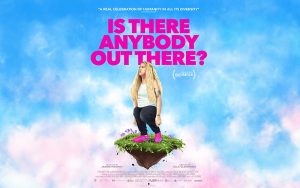 Director Ella Glendening is standing on a floating piece of turf, looking up to her left. She is wearing a light blue t shirt, black trousers and pink shoes. The background is a cloudy sky.