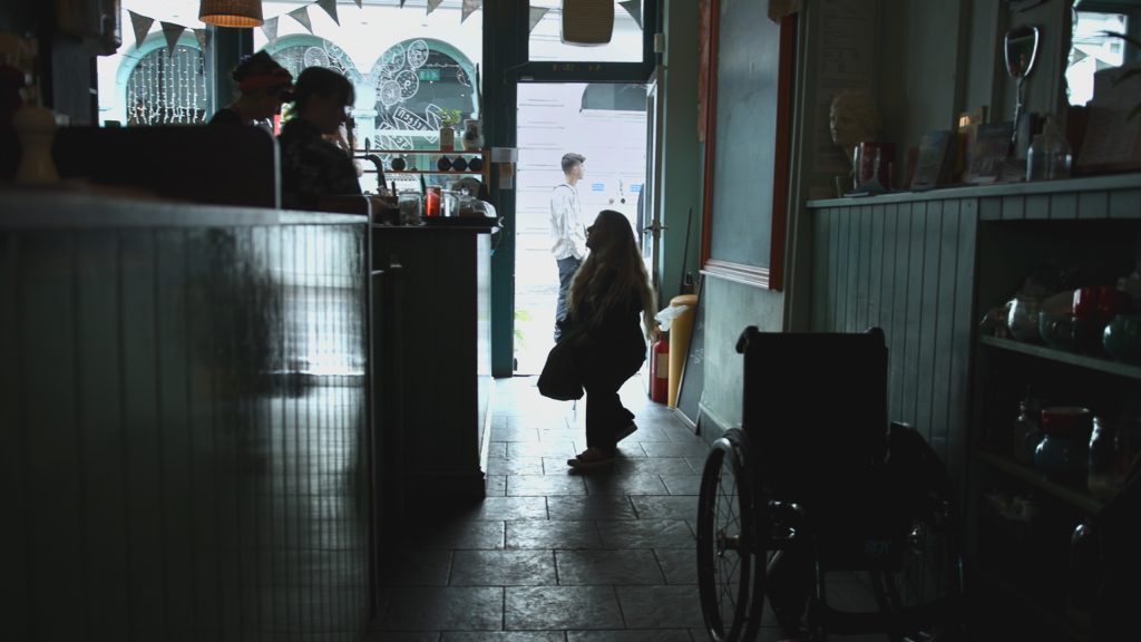 Still from film 'Is There Anybody Out There?' Director Ella Glendining is silhouetted against the front window of a cafe. She is looking at the person behind the counter.