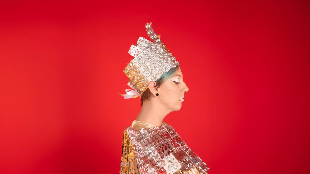 Profile shot of a model wearing an outfit made from empty tablet wrappers. The model is in front of a red background, and is wearing a crown similar to the outfit.