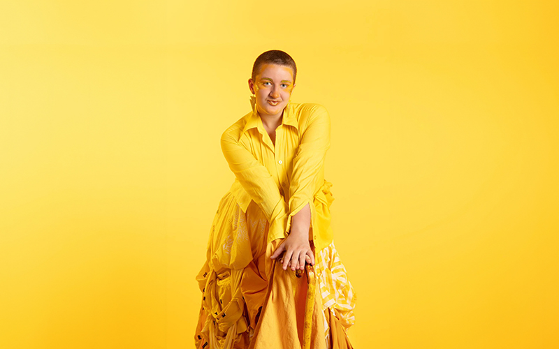 May is sitting on a tall chair in front of a bright yellow background. She is wearing all yellow, looking into the camera with both hands on her knees.