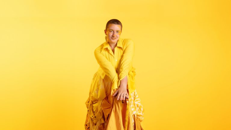 May is sitting on a tall chair in front of a bright yellow background. She is wearing all yellow, looking into the camera with both hands on her knees.