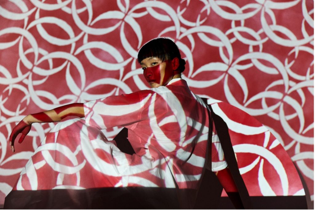 Photography copyright (c) Liberty. The image is of a person looking over their shoulder to address the camera with their left arm raised. A red and white circular and repetitive pattern has been projected onto the person and background, creating an abstract image.