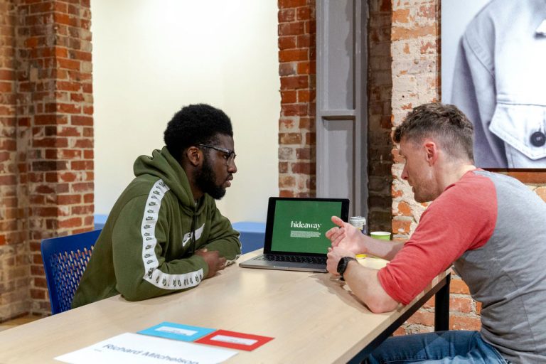 A photo from The Big Book Crit 2024 in Duke Street foyer. The image is of a student and an industry guest sat down at a table reviewing the students work and or portfolio on a laptop.