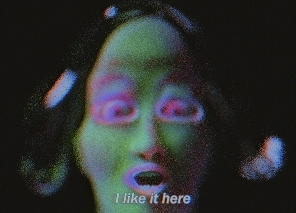 Digital humanoid face with wide eyes, green skin, and pink features, subtitled 'I like it here'