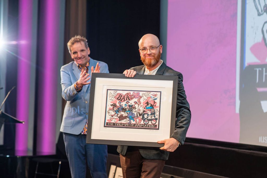 Jon Dunleavy holds a picture in a black frame. He is standing on a stage with a purple curtain behind. A man is standing next to him in a blue shirt.