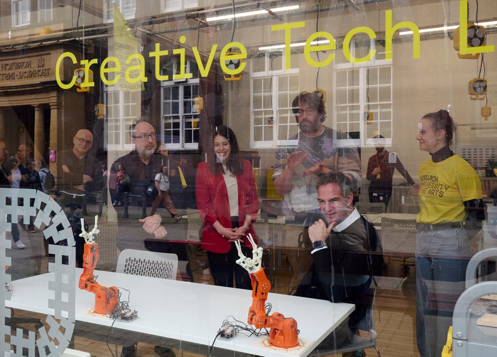 A group of people are looking with interest at orange robot arms. Photo is taken through a window, with lights and reflections across the glass.