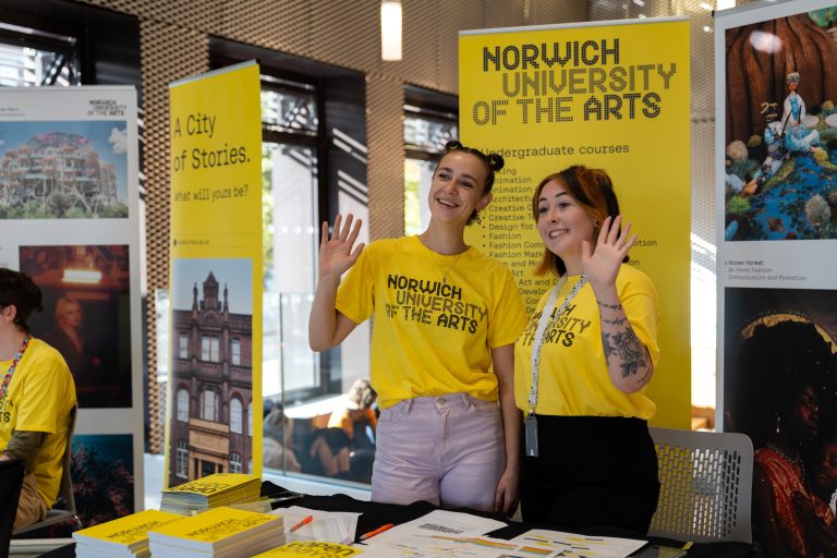 Photograph of student ambassadors waving off camera from behind a desk. They are standing in front of Open day signage.